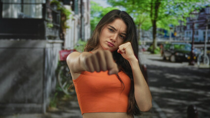 Woman in orange crop top punching with bare fist toward camera on sunlit city street lined with bicycles; self defense determination.