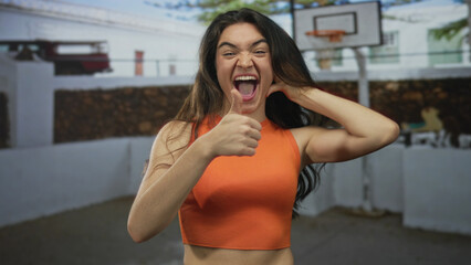 Hispanic teenage woman in orange top makes thumbs up gesture on basketball court beside a building; happiness.