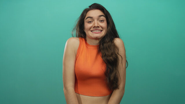 Woman with bare arms and midriff shrugs in a studio wearing an orange crop top and cringing tense shoulders; uncertainty.