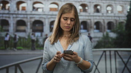 Young blonde woman taps smartphone while standing at ancient coliseum in daylight; concern...