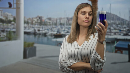 Fototapeta premium Woman holding blue bottle and pointing finger to temple while examining label next to dockside boats at port; curiosity.