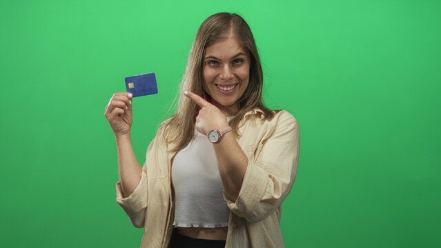 Young blonde woman holds creditcard and points finger to card against green screen in studio set; trust.