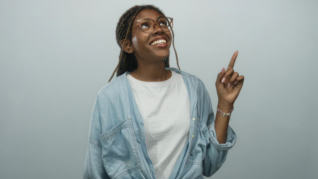 African american woman points finger upward in studio with pale blue backdrop and glasses, wearing denim shirt and white tee; joyful optimism.