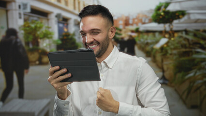 Young man outdoors celebrating success on tablet in urban street setting with a joyful expression and confident posture.