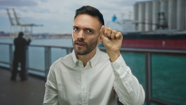 Hispanic man on cruise boat making knocking gesture with ocean and cargo ship in the background under a bright sky.