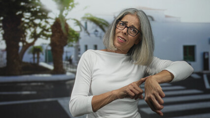Woman points finger to wrist on street crosswalk by white building and palm trees; impatience waiting time.