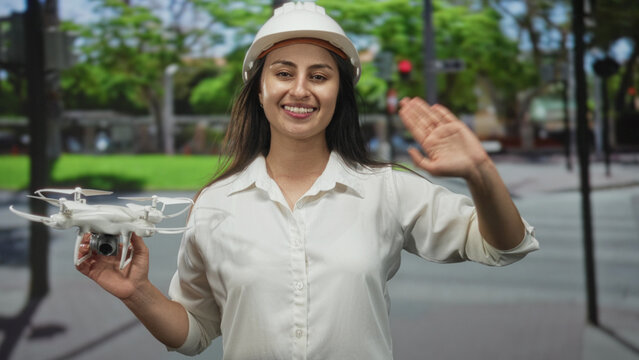 Woman engineer holding drone and waving hand on street wearing hardhat; confidence innovation teamwork.