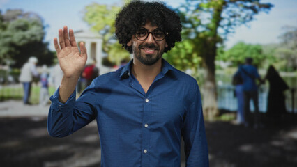 Man with curly hair wearing glasses and blue shirt smiling and raising hand in city park by pond; approachable warmth.