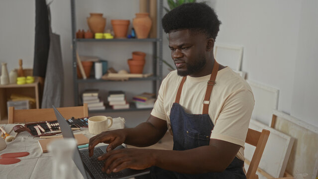 Man typing on laptop with hand on keyboard and holding a ceramic mug at a pottery studio table surrounded by tools and clay pots; creativity focus calm.