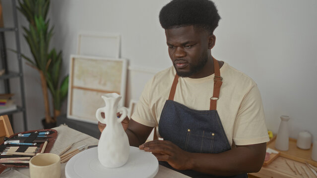 Young man shapes a clay vase with his hands on a pottery wheel in a studio wearing a denim apron and clay tools on the table; concentration. - Powered by Adobe