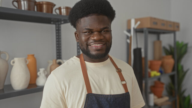 Young african american man wearing apron smiling beside ceramic vases and pottery shelves in studio; pride craftsmanship creativity.
