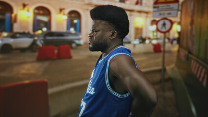 Man hands on hips on city street at night wearing blue basketball jersey, side profile showing muscular arm and strong posture; frustration and solitude.