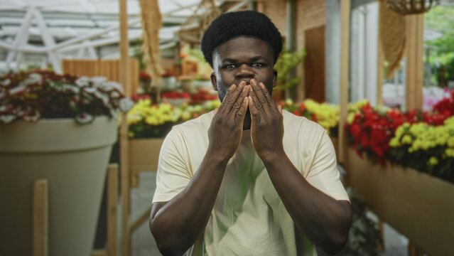 Young african american man clasping hands near colorful potted flowers in building aisle; gentle joy. - Powered by Adobe