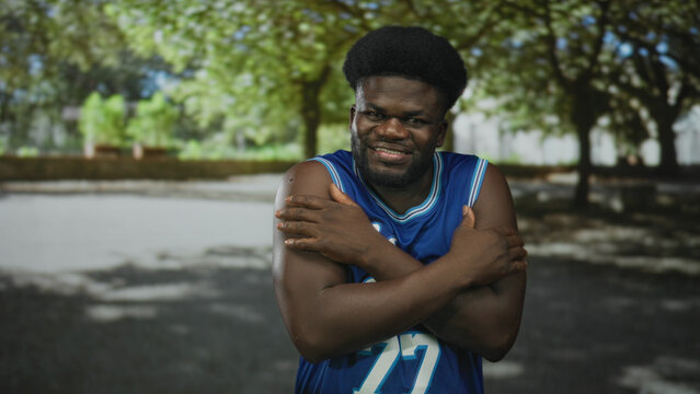 Young african american man in a blue basketball jersey with arms crossed hugging his chest and smiling in a sun dappled forest setting; self comfort contentment.