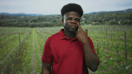 Naklejka premium Man with glasses and beard, finger to cheek near eye in forest vineyard rows wearing red polo; pensive reflection.