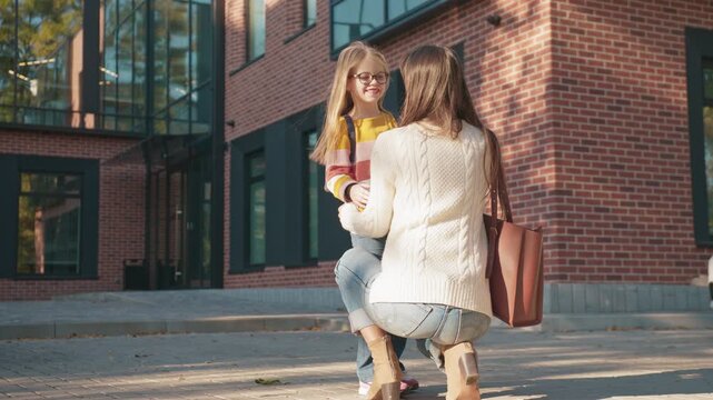 Schoolgirl running toward female parent with arms open on brick walkway. Child carrying backpack rushing for embrace. Woman kneeling waiting to hug daughter near modern campus entrance.