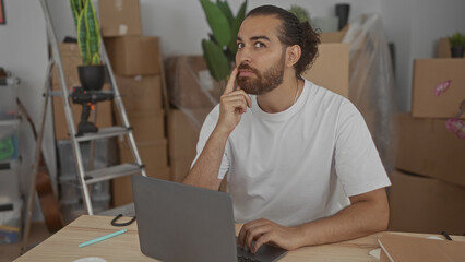 Man with beard and bun, hand on chin, typing on laptop at table surrounded by moving boxes and ladder in building; focused concentration.