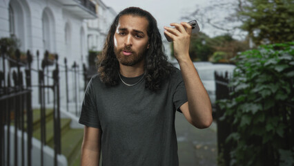 Young man holds smartphone to ear on city street near iron railings and white townhouses wearing dark tee; urban frustration.