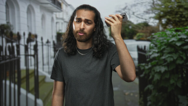 Young man holds smartphone to ear on city street near iron railings and white townhouses wearing dark tee; urban frustration.