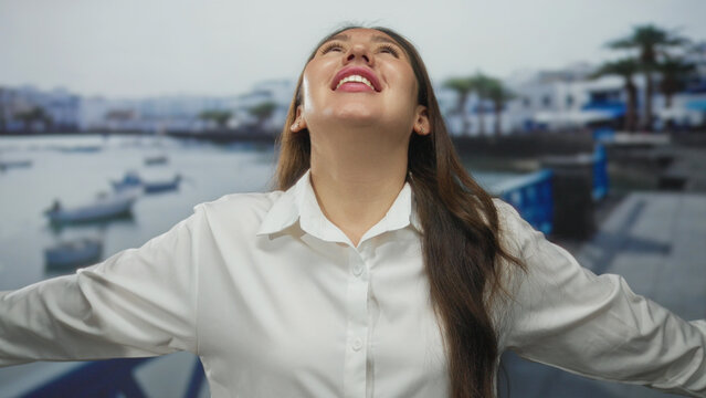 Young hispanic woman stands with arms wide and closed eyes beside a docked boat at sunlit seaside port pier; serenity. - Powered by Adobe