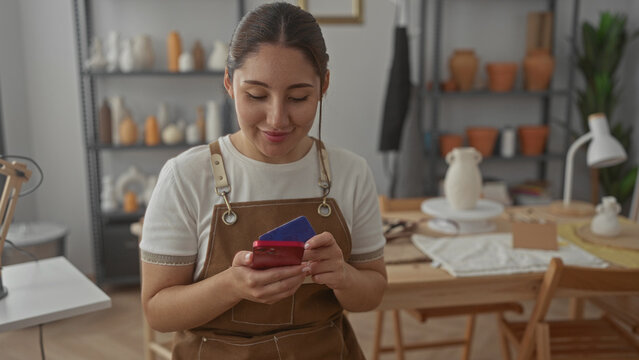 Woman in brown apron taps smartphone screen holding a blue creditcard in studio workshop with pottery shelves; creative satisfaction.