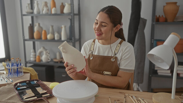 Young hispanic artisan woman holds ceramic vase with hands in studio surrounded by pottery shelves and sculpting tools; serenity. - Powered by Adobe