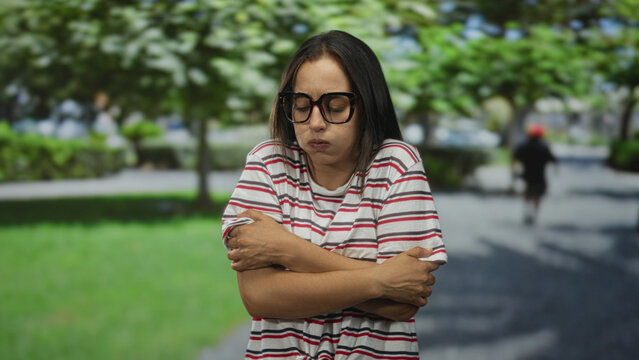 Woman in striped shirt and glasses hugging herself with arms crossed over chest on a park pathway under green trees; cold discomfort. - Powered by Adobe