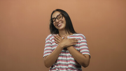 Woman with hands crossed over chest wearing glasses and a striped shirt, smiling with closed eyes in peach studio; gentle gratitude.