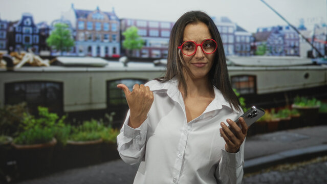 Woman in white shirt holding smartphone and thumb pointing to side on a street with canal boat and buildings; skepticism. - Powered by Adobe