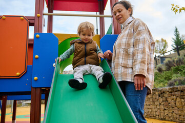 Mother Helps Happy Child On Green Slide At Outdoor Playground On Autumn Day