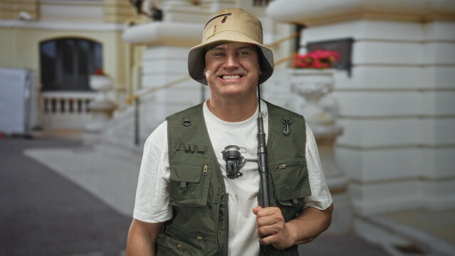 Man holding fishing rod with bare hand, smiling in front of building, wearing fishing vest and bucket hat; leisure contentment.