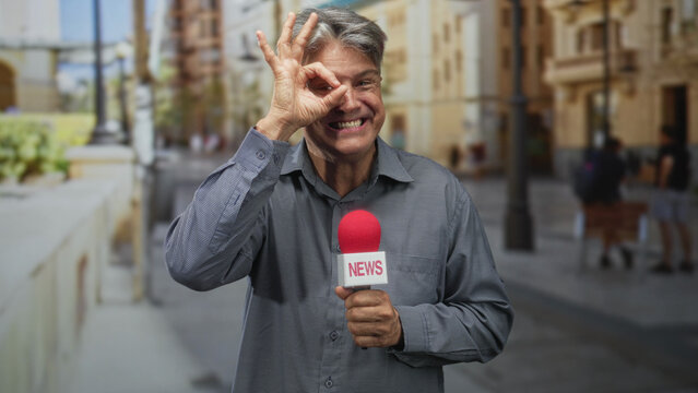 Man holding a red news microphone, smiling, and making an ok sign with fingers over his right eye while reporting on a sunny street; playful friendliness.
