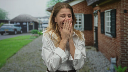 Woman covering mouth with hand at building entrance outside home, smiling and laughing in doorway while wearing white shirt; surprise welcome.