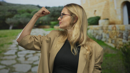 Woman with glasses flexing arm in strength gesture outdoors in a sunny park wearing beige blazer...
