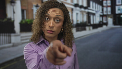 Hispanic woman pointing finger on city street in afternoon light with serious stare and tense stance; assertion defiance confidence resistance.