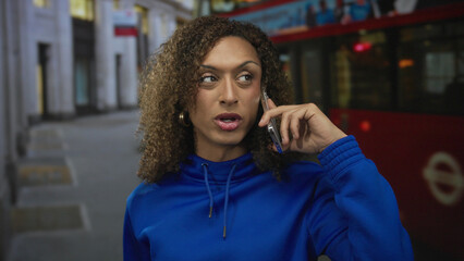 Fototapeta premium Woman wearing blue hoodie holding smartphone to ear on city street with red bus behind; isolation reflection.