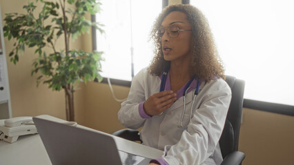 Curly haired woman doctor holding stethoscope holds hand on chest at clinic desk during indoor...