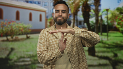 Man making timeout gesture with hands, standing in a green park with palm trees and a white building behind him; confidence.