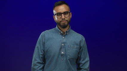 Obraz premium Young hispanic man wearing denim shirt with glasses flexes arm and shows bicep in blue studio; confidence strength.