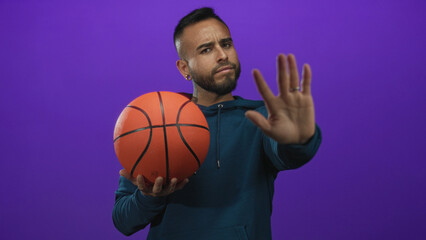 Young man holding basketball with outstretched hand stop gesture in purple studio; determination focus.