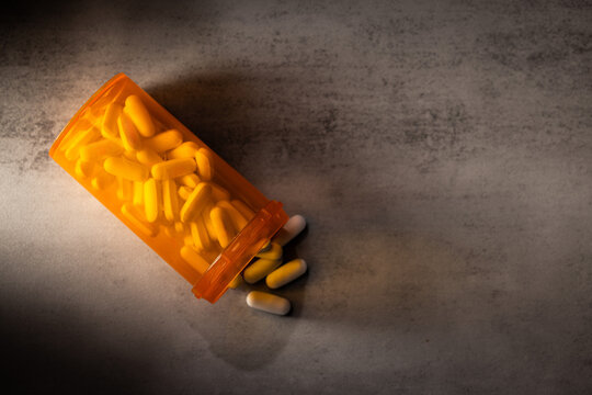 A dramatic, low-key photograph of capsules and pills spilling from an orange bottle onto a concrete surface, highlighted by hard light and deep shadow.