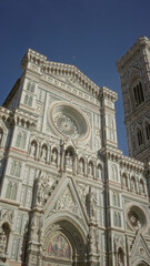 Fototapeta premium Campanile and cathedral facade standing tall with detailed green white and pink marble ornament, rose window and statues on a building in florence italy; awe.