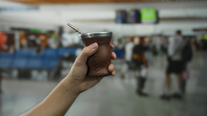 Man holding mate cup in hand at busy airport terminal with blurred background of travelers and seating, illustrating a casual travel ambiance.