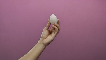 Caucasian man's hand gracefully holds a beauty blender sponge against a vibrant pink background, emphasizing skincare and cosmetics in an isolated setting.