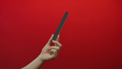 Man hand holding nail file against red wall background showcasing isolated manicure tool with notable grip technique and focus on grooming implement texture.