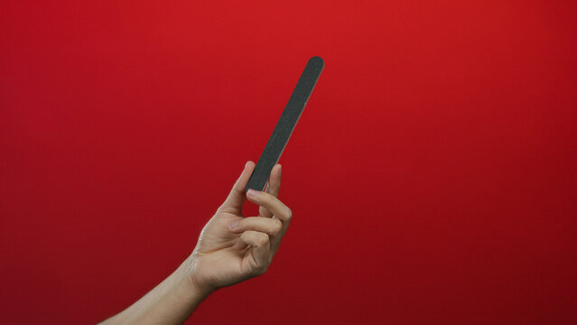 Man hand holding nail file against red wall background showcasing isolated manicure tool with notable grip technique and focus on grooming implement texture.