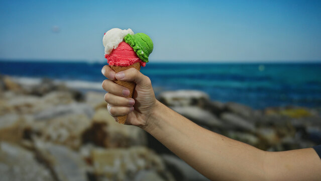 Man holding colorful ice cream cone on beach against clear blue sky and ocean backdrop showcasing serene seaside relaxation. - Powered by Adobe
