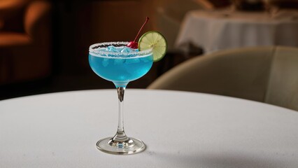 A blue cocktail drink garnished with a cherry and lime slice on a white table with a blurred caf&Atilde;&copy; background.