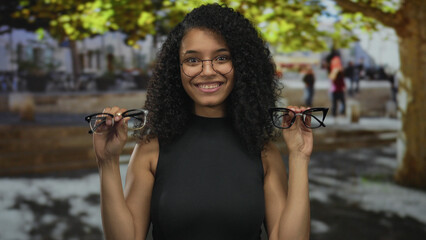 Woman smiling outdoors holding eyeglasses in a vibrant city setting with trees and people walking...