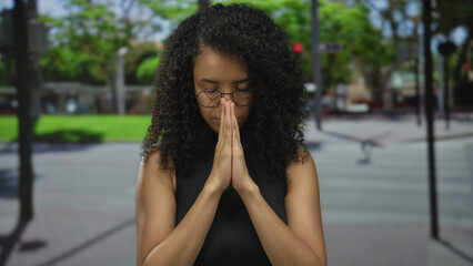 Young woman praying outdoors on a city street with blurred green background, eyes closed, hands together, expressing peace and tranquility, wearing black sleeveless top and glasses.